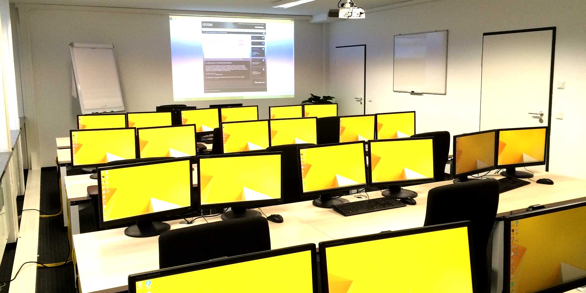 Schulungsraum mit Tischen, Bildschirmen, Leinwand und Beamer
