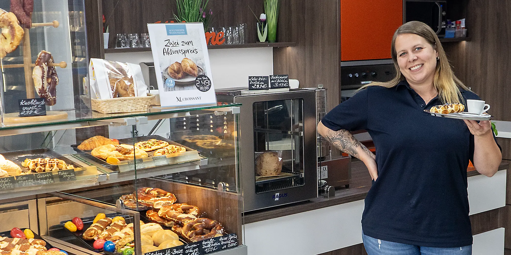 Mitarbeiterin steht im Bistro und präsentiert lächelnd einen Teller mit Kaffeetasse